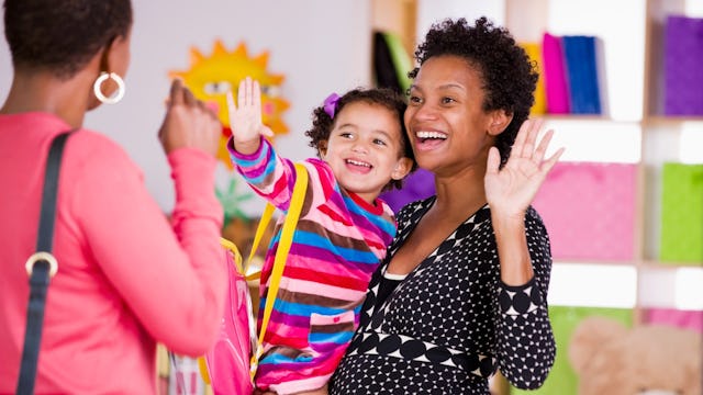 A happy and smiling mother holding a child in her arms as they're waving at a preschool teacher