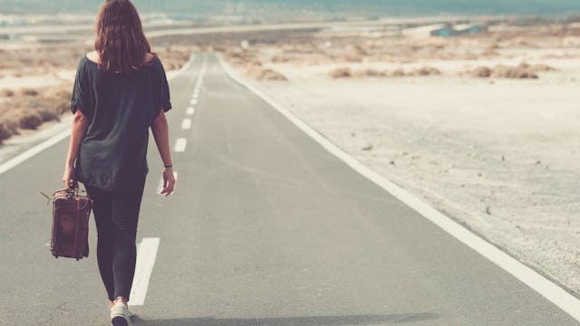 A woman in a black shirt and black trousers carrying a brown suitcase walking on the empty road with...