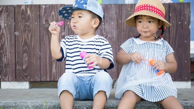 A boy in a blue-white striped T-shirt, blue denim jeans, and a cap blowing bubbles next to a girl in...