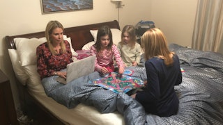 A mom sitting on the bed and working on her laptop while her daughters play a board game beside her