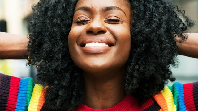 A black woman with afro haircut wearing a black and rainbow-colored sweater and smiling with eyes cl...