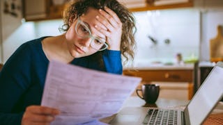 A woman at the table next to her laptop looking over her bills and holding her head