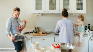 A husband in a blue shirt holding a newborn and looking at a newspaper, a wife cooking with a toddle...
