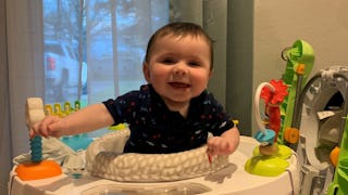 A baby boy sitting in a white chair surrounded by his toys and smiling