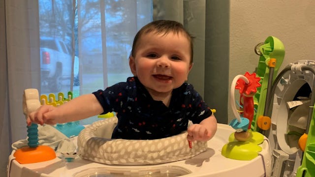 A baby boy sitting in a white chair surrounded by his toys and smiling