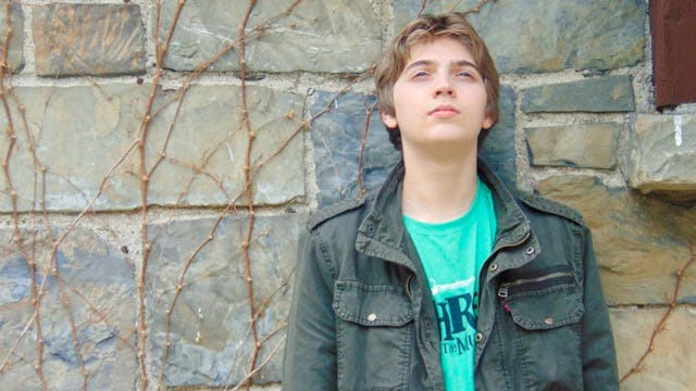 16-year-old boy in a green shirt and dark-olive jacket leaning on the wall and looking up