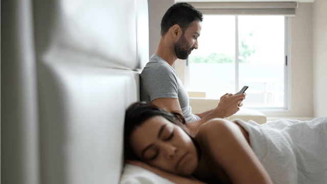 A woman lying asleep in bed while her husband sits behind her, looking at porn on his phone.