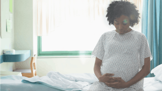 A pregnant black woman sitting on a hospital bed with her hands on her belly while wearing a hospita...