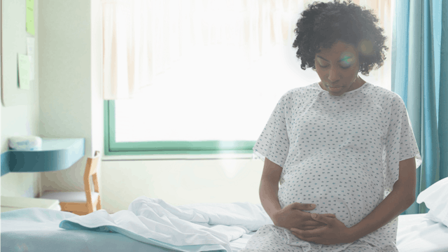 A pregnant black woman sitting on a hospital bed with her hands on her belly while wearing a hospita...