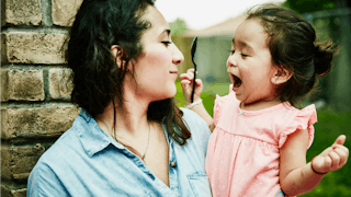A mom in blue denim shirt looking at her daughter dressed in a pink dress with a spoon while holding...