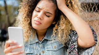 A girl with an afro hairstyle looking at an MLM message on her phone with a worried facial expressio...