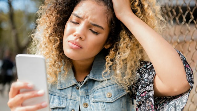 A girl with an afro hairstyle looking at an MLM message on her phone with a worried facial expressio...