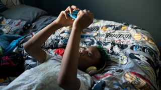 An autistic tween in a grey T-shirt wearing headphones while playing with his phone in a bed