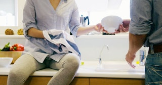 Woman asking husband for help with washing the dishes. She has a kitchen towel in one hand and is ta...