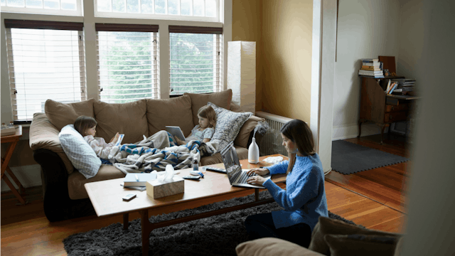 A mother wearing a blue shirt sitting on the ground and her two kids lying on the couch near a table...