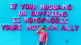 Man standing in front of a blue wall with pink "if your husband or boyfriend is homophobic you're no...