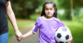 A girl in a purple shirt holding a soccer ball walking and holding her soccer mom's hand