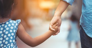 A parent in a blue shirt and denim jeans and kid in a blue-white polka dot dress holding hands while...