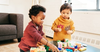 A young child in a burgundy sweater next to a child in a mustard-yellow sweater assembling blocks on...