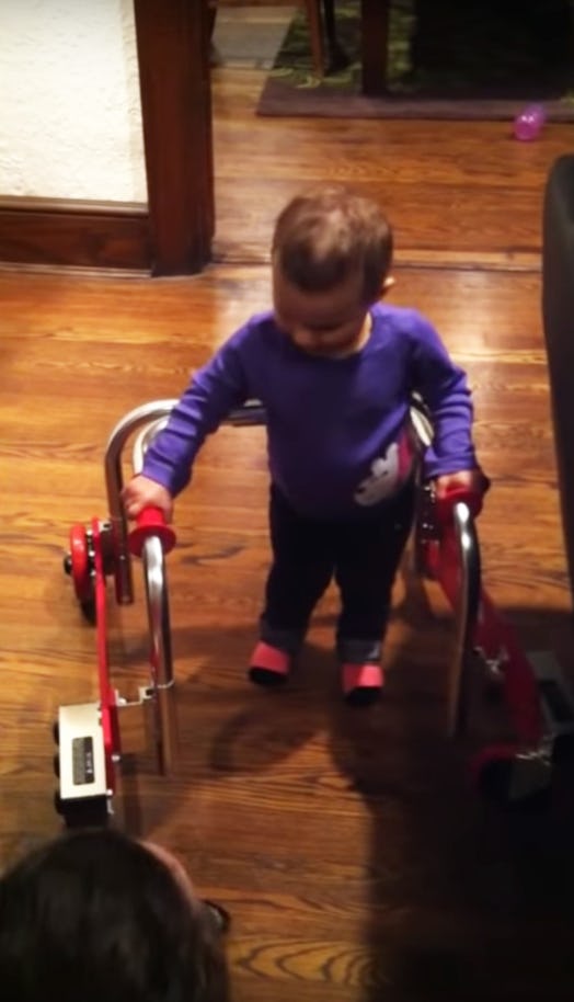 A toddler girl wearing a purple shirt and black pants using a walker that is helping her learn to wa...