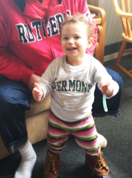 A toddler girl wearing a white shirt with "Vermont" on it while smiling and someone behind her weari...