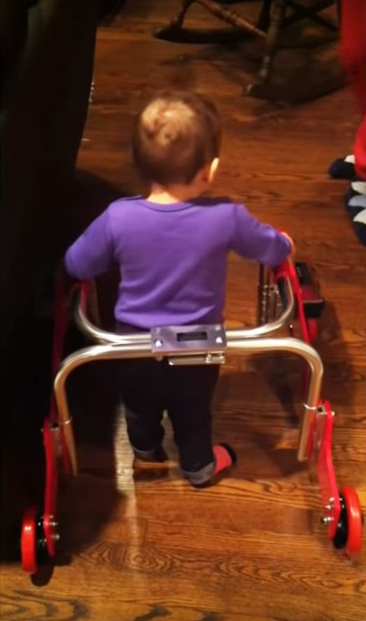 A toddler girl wearing a purple shirt and black pants using a walker that is helping her learn to wa...