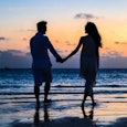 Married couple walking down on a beach holding hands while Sun is going down.