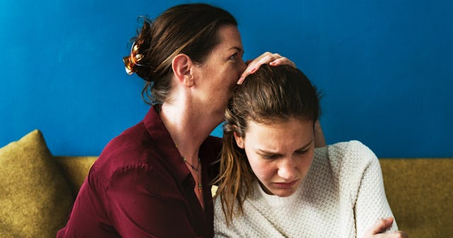A worried mom in a red shirt is kissing her child who is suffering from addiction while they are sit...