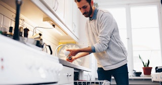 A man wearing a blue shirt, white sweater and jeans putting dishes in the dishwasher