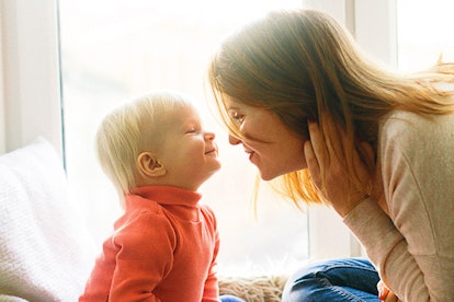 A brunette woman in a beige sweater holding her hair and leaning towards her blonde baby wearing an ...