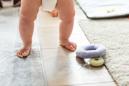 A baby in a diaper walking over beige tiles and grey carpet and a round grey plush baby toy on the f...