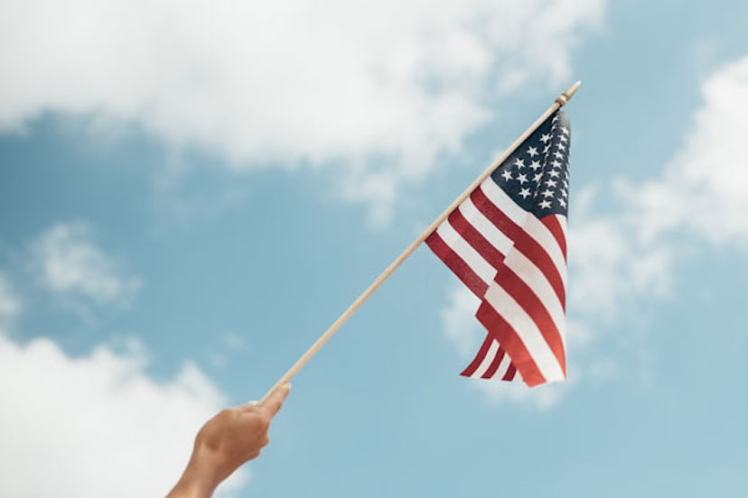 A hand waving an American flag on a wooden stick with the blue sky and clouds behind it