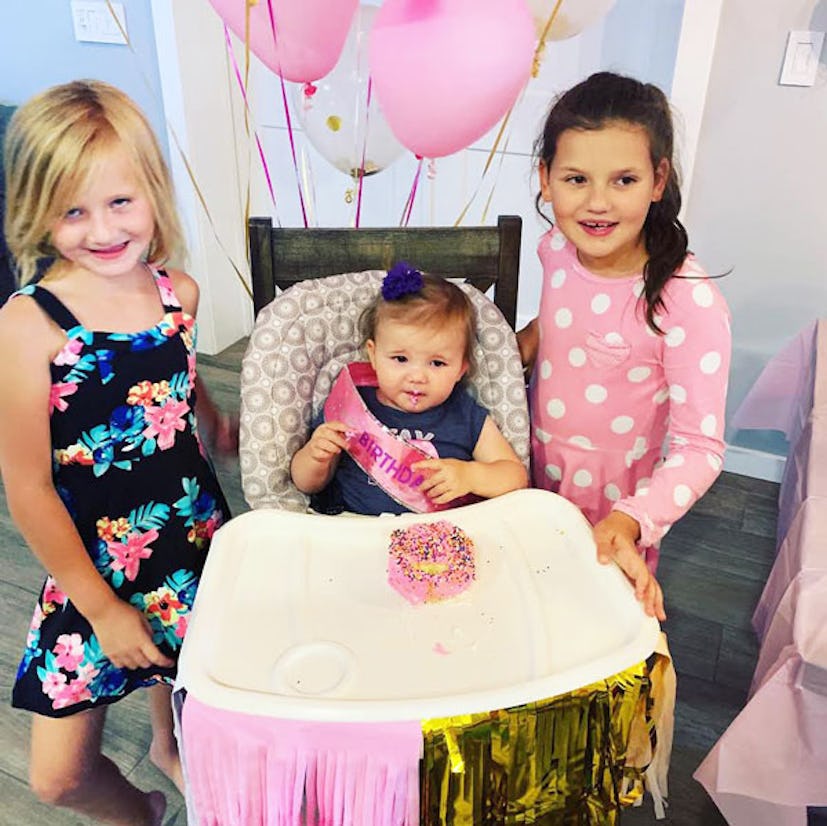 Birthday girl toddler with her two sisters at her party with a pink cake and balloons