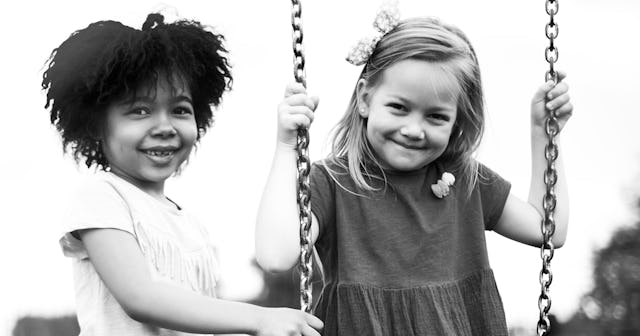 A light-haired girl is sitting on a swing and next to her is her friend with short curly hair in bla...