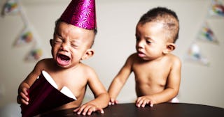 One twin wearing a purple birthday hat and crying, while the other twin is looking at him