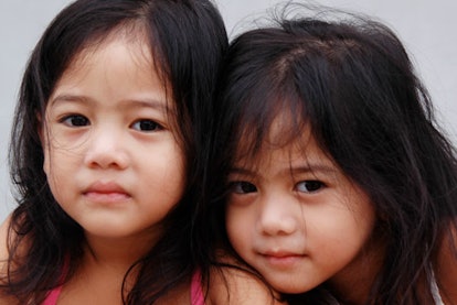 Twin girls with long black hair looking at the camera while one is leaning on the other 