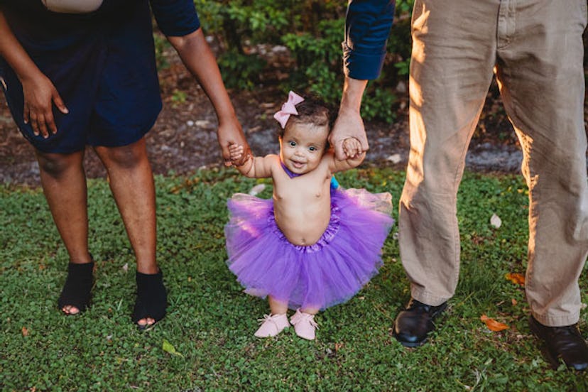 A little girl in a purple ballet skirt holding her parents arms.