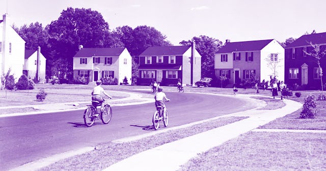 Two children riding their bikes down a street with rows of houses on both sides in a village all blu...
