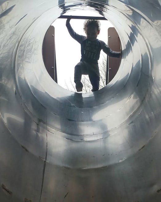 A curly-haired toddler on the top of an open metal tunnel segment for a playground
