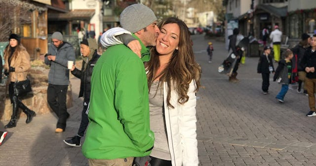 A gentleman in a green jacket and gray cap affectionately pecking a woman donned in a white jacket o...