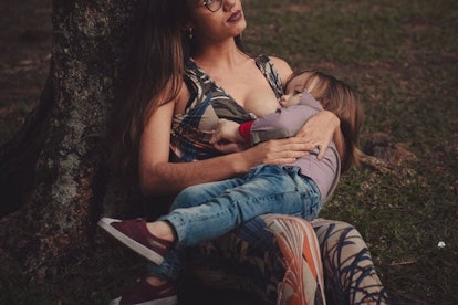 A mother breastfeeding her toddler, leaning on a tree