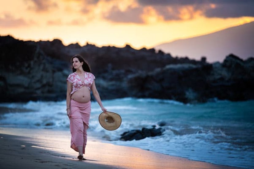 A pregnant woman walking next to the sea in a pink top and skirt while holding a straw hat
