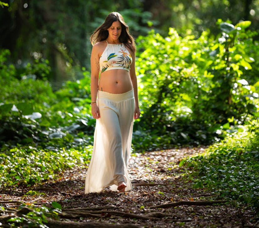 A black-haired pregnant woman walking through a forest in a white top and skirt 