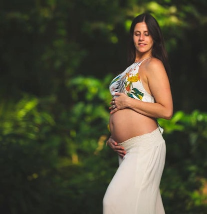 A pregnant woman with her hands on her baby bump while posing in a beach white top and skirt 