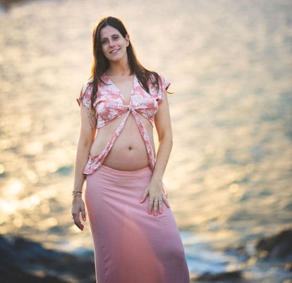 A black-haired pregnant woman standing next to the sea in a pink top and skirt
