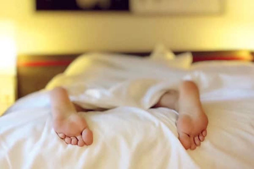 A person sleeping on their stomach on a white bed sheet with only the feet sticking out under the sh...