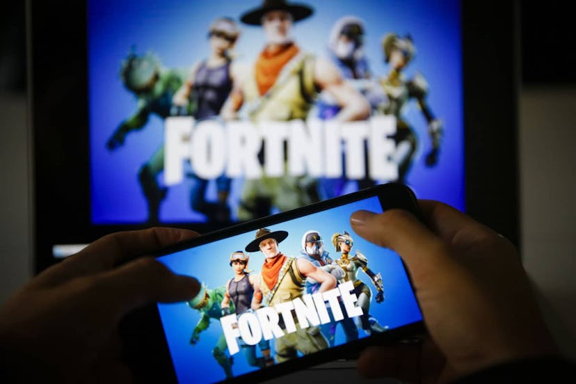 A child playing Fortnite on his smartphone in the dark with Fortnite on the TV in the back slightly ...