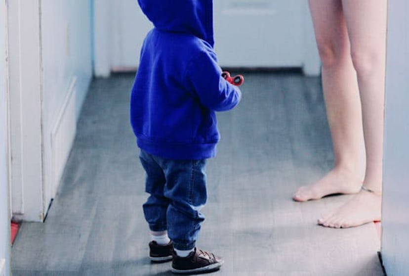 A toddler boy in a blue hoodie, blue denim jeans and brown shoes standing next to his mother
