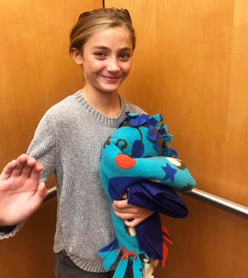 Sara Gibney's daughter in a gray sweater waving with and elevator holding a blue patterned blanket i...