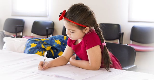 A brunette girl with a long braid in a burgundy shirt and headband with a flower writing something d...
